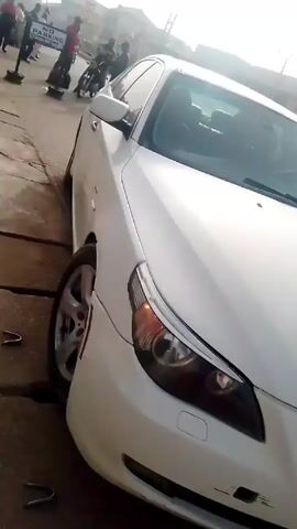 The video shows a white BMW sedan parked on a street. The camera pans along the side of the car, showing the tires and the car's features. A man wearing a red t-shirt, jeans, and a black crossbody bag walks past the car. A white motorcycle is parked nearby. The environment appears to be a street in a town or city, with buildings and other people visible in the background.