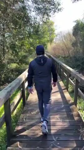 A man walks away from the viewer along a wooden bridge with railings. The man is wearing a dark jacket with a hood, dark pants, a baseball cap, and white sneakers. The bridge is surrounded by trees and brush. The weather is sunny.