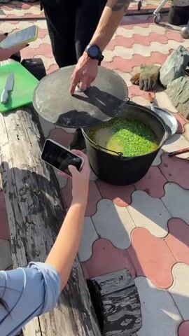 Two individuals are present. One, wearing black pants and sneakers, is holding a large black pot with a lid. The other, in a blue shirt, is holding a smartphone in landscape orientation, recording. The pot contains a yellow broth with what appears to be chopped green vegetables and some orange pieces, possibly carrots. The scene takes place outdoors on a tiled patio with a patterned arrangement of pink and white interlocking tiles. Nearby are large rocks, a metal grill with a fire, two black pots on the grill, a metal ladle, a slotted spoon, work gloves, and a green cutting board. Wooden logs are also visible. The overall impression is of an outdoor cooking event.