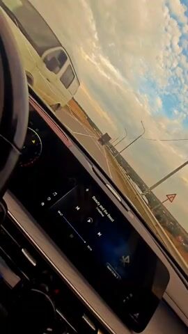 The video shows a view from the interior of a car, looking out at a road and the sky. The car's dashboard is visible, with a large touchscreen display showing a music player interface. The song playing is titled "Beauty and the Beast." The car appears to be in motion, as the scenery outside is blurred and the perspective is angled. The sky is cloudy, with sunlight breaking through, suggesting it is either sunrise or sunset. The road ahead is straight and extends into the distance, with some utility poles lining the sides. There is a truck in the distance on the road.