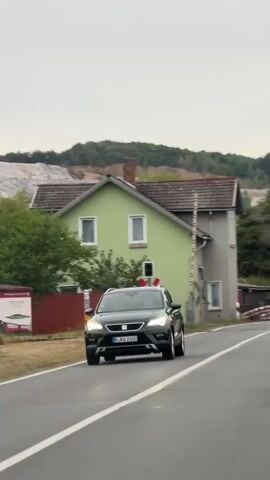 A car drives past a railroad crossing signal and a house. The signal is red and white, with a red light at the top. The house is light green and gray, with a red chimney. There are trees and a hill in the background. The sky is overcast.