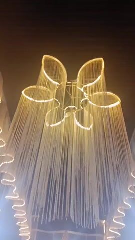 The video shows an ornate chandelier with multiple circular tiers adorned with hanging strands of small, light-colored beads. The chandelier is illuminated by warm, golden lights, creating a sparkling effect. Below the large chandelier, several smaller chandeliers are visible, along with a wavy, illuminated decorative element that frames the scene from the sides. In the background, a gathering of people is visible, some seated and others standing, suggesting an event or celebration. The overall atmosphere appears festive and elegant, with a focus on the elaborate lighting and decor.