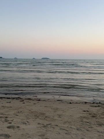 The video presents a serene panoramic view of a sandy beach and the ocean during either sunrise or sunset. The sky displays a soft gradient of light blue transitioning to faint pink and orange hues, indicating the low position of the sun. Gentle waves rhythmically lap against the shore, creating a tranquil soundscape.

The scene begins with a wide shot of the ocean, where several small, dark islands or landmasses are visible on the distant horizon. The foreground shows a clean sandy beach with some natural debris, such as seaweed and small twigs, scattered near the water's edge.

As the camera slowly pans to the left, more of the expansive ocean comes into view. A solitary sailboat, along with a few other smaller boats, can be seen anchored or sailing calmly in the distance. The pan continues, revealing a picturesque coastline characterized by lush, dark mountains and hills that stretch along the horizon, framing the beach. The sand extends continuously along the shoreline.

The overall impression is one of peaceful natural beauty. There are no people visible on the beach or in the water, contributing to the tranquil atmosphere. The setting is a quiet, untouched beach, likely located in Baan Klong Prao, Thailand, given the provided context. No text is visible in the video.