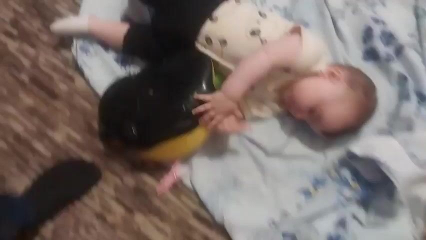 A baby is lying on its back on a blue patterned blanket next to a black and yellow penguin toy. The baby is wearing a light-colored onesie with panda bear prints and black pants with white socks. The baby is holding onto the penguin toy with both hands, and its head is tilted to the side. The baby appears to be drowsy or asleep. The environment is indoors, with a wooden floor visible in the background.