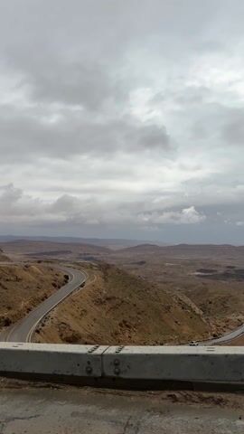 The video shows a vast, arid landscape under a cloudy, overcast sky. The terrain is predominantly brown and barren, punctuated by gentle hills and valleys. A winding asphalt road cuts through the landscape, disappearing into the distance. A gray guardrail stands in the foreground, slightly wet, suggesting recent or ongoing rain. A vehicle is visible on the road in the distance. The sky is dominated by gray clouds, obscuring the sunlight and contributing to the overall somber atmosphere. The camera appears to be stationary, capturing the scene from an elevated viewpoint.