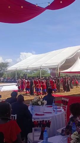 The video shows a group of people performing a traditional dance in front of an audience seated at tables under a white tent. The dancers are dressed in red and black clothing, and they are moving in a synchronized fashion. The audience is watching attentively. There is red draping above, and a crowd of people visible further in the distance. The tables are set with white tablecloths, plates, glasses, and water bottles. There are also floral centerpieces on the tables. The environment appears to be outdoors, with a clear blue sky and a few scattered clouds. The ground is dirt or sand.
