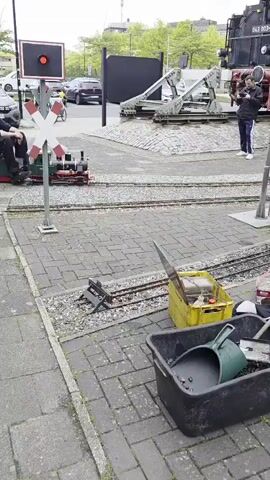 The video depicts a miniature train setup in an outdoor environment, possibly a park or recreational area. Several people are riding on small, open-top train cars being pulled by miniature locomotives. The locomotives are different colors including green and blue. The train is traveling on a narrow-gauge track that runs along a brick or paved path. There are other stationary train cars on adjacent tracks. A train station signal with a red light and X-shaped cross buck is visible, along with what appears to be a larger, stationary locomotive in the background. People can be seen filming the miniature train as it passes. Several containers near the tracks hold tools, including a shovel and coal. The environment includes trees, grass, and buildings in the distance.