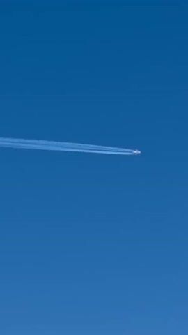 The video shows a jet airplane high in a clear blue sky. The airplane is leaving a long white contrail behind it. The plane is moving from left to right, away from the camera.