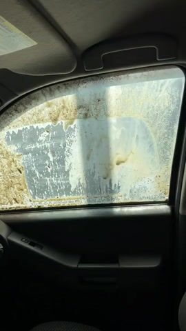 The video shows a close-up view from inside a car, focusing on the driver's side window. The window is heavily covered in dirt and grime, obscuring the view outside. The car's interior, including the door panel, handle, and window controls, is visible. The sunshade above the window is also present. There is no visible action or interaction occurring within the frame.