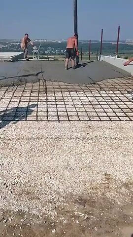 A concrete pump hose is being used to pour wet concrete onto a grid of rebar that is laid out on a surface. Three men are visible. One man in the foreground on the right is wearing a light-colored shirt and dark shorts and is standing near the edge of the pouring area, facing away from the camera. A second man, shirtless and wearing dark shorts, is operating the concrete pump hose, directing the flow of concrete. A third man, also shirtless and wearing light-colored shorts, is standing further back on the left, appearing to be supervising or assisting. The environment is outdoors, with a clear blue sky and a distant landscape of buildings and trees. The ground in the foreground is covered with small, light-colored gravel.