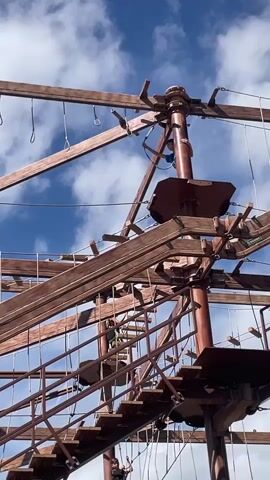 A low-angle medium shot captures an outdoor ropes course against a bright blue sky with scattered white clouds.  The ropes course is constructed from thick wooden beams and planks, forming multiple levels of walkways, stairs, and platforms.  Several people are visible on the structure, all wearing safety harnesses.  In the upper portion of the frame, a person in a blue shirt and harness is positioned on a platform, appearing to be moving along a cable.  Below, on a set of wooden stairs, another person in a dark shirt and harness is descending.  Further down, near the bottom of the frame, a third person in a blue shirt and harness is ascending the stairs.  Rope bridges and netting crisscross between the wooden elements, indicating various pathways within the course.  The overall impression is of an active recreational environment.