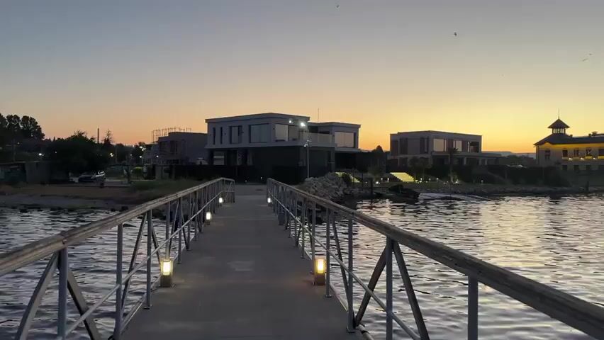 A video recorded at dusk shows a pier extending into a body of water. Modern houses are visible on the shore in the background, and a large, older building with a tower is illuminated. The sky displays a gradient of colors from orange and pink near the horizon to deep blue overhead, with a few birds flying in the distance. The water reflects the sky and the lights from the pier and the buildings.
