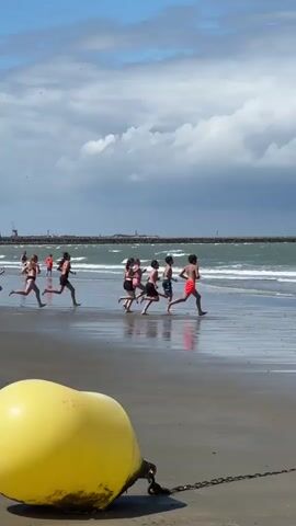 A group of children are running along a beach towards the ocean. They are wearing swimsuits and appear to be playing or racing. The water is calm with small waves. In the background, there is a long pier with a lighthouse at the end, and several sailboats can be seen on the horizon. The sky is partly cloudy with a mix of sun and dark clouds. In the foreground, a large yellow buoy is partially visible.
