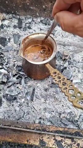 A person's hand is stirring coffee in a copper pot over hot coals. The coffee is dark brown and frothy. The pot has an ornate brass handle. The coals are gray and black, with some embers glowing. The background is out of focus but appears to be an outdoor setting.