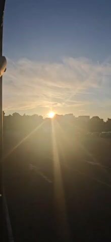 The video shows a view of a car storage lot during sunrise. The sun is low in the sky, casting long rays of light across the scene. In the foreground, there is a paved road with white markings. Beyond the road, a vast lot filled with rows of cars is visible. The cars are parked close together, and appear to be new or in good condition. In the background, there are trees and some buildings. The sky is blue with scattered clouds. The overall impression is one of a quiet and orderly scene, with the natural beauty of the sunrise adding a touch of warmth and color.