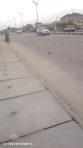 This media file captures a street scene in Adesanya, Nigeria. The primary subjects are a person on a motorcycle and several cars driving on a paved road. The setting is an urban or semi-urban environment with buildings visible in the background. The activity appears to be ordinary street traffic.

Notable details include the time of day, which seems to be daytime given the lighting, and the weather appears overcast. The presence of streetlights suggests it might be dusk or an area that uses them during the day. The road is wide and has a concrete sidewalk along the side. There is a small pile of debris on the sidewalk. The overall mood is neutral, depicting a typical street scene.

Visible text in the media file includes the watermark "TECNO SPARK 10c" at the bottom left corner. There is also a logo with a "Z" shape and some text on a white sign in the background on the left side of the road, but the text is too blurry to read.