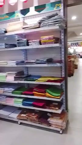 The video pans across a brightly lit retail store filled with shelves stocked with goods. The shelves are organized and display a variety of items, appearing to be fabric or paper products based on their stacked and folded presentation. The color palette of the products ranges from pastel shades to vibrant hues. In the foreground, a person wearing a light-colored hijab and a pink top walks towards the camera, carrying a dark bag. As the video progresses, other individuals are visible, including a child in a red outfit running through the aisles, a woman in a light grey hoodie and black skirt, and another person in a beige hijab. The environment suggests a shop selling textiles, crafts, or stationery, with promotional banners hanging from the ceiling. The floor is a light-colored tile or laminate.