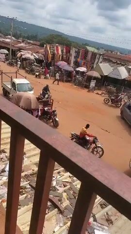 The video shows a view from an elevated position, likely a balcony or bridge, overlooking a bustling street in what appears to be a market town. The street is unpaved, with a reddish-brown hue. There are numerous motorcycles, some with riders, moving along the street. A small white truck is parked in the middle of the road, with what looks like a motorcycle and a covered trailer attached to it. The sides of the street are lined with makeshift stalls and shops, many covered with umbrellas or awnings. There are various fabrics displayed, suggesting some stalls may sell textiles. People are walking around, presumably vendors and shoppers. In the background, there is a landscape of hills covered with green vegetation, under a cloudy sky. The video is taken from behind a metal railing painted a reddish-brown color. There is some trash visible on the ground near the railing. The video appears to be shot with a mobile device.