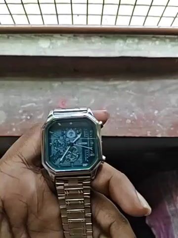 A close-up shot shows a person holding a silver-banded watch with a square, octagonal-shaped face. The watch face is a dark blue-green color and appears to have a complex mechanical design. The person's hand is in focus, with the watch positioned in the palm and fingers. The background is blurred, but suggests an outdoor or industrial setting with a concrete or stone surface and a metal grate. The lighting is somewhat dim, casting shadows on the hand and watch.