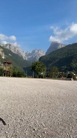 The video captures a landscape scene on a bright, sunny day with a clear blue sky dotted with fluffy white clouds. In the foreground, a wide expanse of gravel occupies the lower portion of the frame. Beyond the gravel, a line of trees separates the viewer from a small town nestled at the base of a lush, green mountain. The town features buildings with orange and brown roofs, interspersed with more trees. The mountain itself is covered in dense vegetation, and its rocky, jagged peaks rise majestically in the distance, shrouded in a light haze. On the far left, a wooden observation tower with a red roof is visible. Some indistinct figures can be seen walking in the middle ground of the scene.
