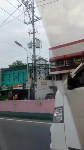 The video captures an urban street scene in Pasay, Philippines, as viewed from a moving vehicle. The sky is overcast throughout, suggesting cloudy weather.

The scene features a pink road barrier running parallel to the direction of travel, separating the roadway from the buildings and sidewalks. Dominant features are numerous utility poles heavily laden with a dense network of overhead wires, characteristic of urban infrastructure.

Various buildings line the street. At the beginning, a two-story green building with commercial spaces on the ground floor is visible; a sign for "PAGASA" is partially discernible. As the vehicle proceeds, more buildings appear, including one with dark glass windows and another multi-story, light yellow building that has scaffolding on one side, suggesting ongoing construction or maintenance.

Palm trees are scattered along the roadside, adding a touch of nature to the urban environment. Several vehicles are seen, including white vans (one with "THE RIGGING CO." partially visible on its side), other cars, and a scooter. A white Toyota Innova is prominently seen in the foreground towards the end of the video. People are briefly visible walking on the sidewalk at one point. The video depicts a typical daytime drive through a bustling urban area.