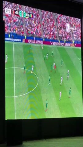 The video shows a large screen displaying a soccer match. The score is Belgium 0, Senegal 0. The time is 02:14. The players are wearing green and white uniforms. The stadium is full of spectators. The screen also displays advertisements for Qatar Airways and World's Best Airline. The camera is positioned at an angle, looking up at the screen.