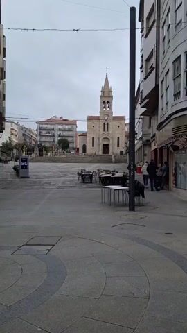 The video shows a town square on an overcast day, with a church in the background. The church is made of light-colored stone and has a steeple with a cross on top. There's a clock on the front of the church tower. In the square, there are tables and chairs, likely for outdoor dining. A few people are standing near a building on the right side of the frame, appearing to be conversing. Some other people are walking across the square towards the church. The ground is paved with a mix of gray stone, and there are circular patterns in the paving near the camera. A tall, dark pole stands near the right side of the frame. At one point, a finger briefly obstructs the view.
