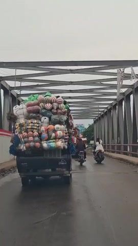 The video shows the rear view of a black pickup truck heavily overloaded with various goods. The truck is driving on a bridge with a metal truss structure overhead. The load consists of many large, round objects with stripes, likely balls or inflated items, stacked high. There are also bags of goods, possibly textiles or produce, bundled together. Attached to the side of the truck's load are strings of colorful, decorative items. In front of the truck, two motorcycles are visible, also in motion on the bridge. The road surface appears wet, suggesting it may have rained recently or the weather is overcast. The overall environment suggests a busy road or transportation route.