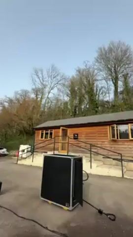 The video shows a black box with lights on it. The box is in front of a one-story wooden building with a few windows. Trees are in the background. The sky is blue. The box shoots out five streams of fire.