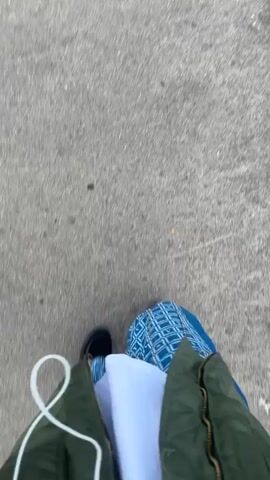The video shows a first-person perspective of someone walking on a paved surface, likely a sidewalk. The person is wearing blue jeans with a patterned design, black shoes, and a green jacket over a white shirt. The camera is pointed downwards, focusing on the person's legs and feet as they move. The ground appears to be a mixture of asphalt and concrete, with some pebbles and debris visible. The lighting suggests it is daytime. The movement of the camera indicates the person is walking forward at a steady pace. There are no other people or objects clearly visible in the frame besides the person walking and the ground.