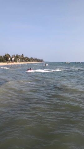 The video shows a tropical beach scene on a sunny day. In the foreground, the ocean is slightly choppy, with gentle waves. A person is riding a red and white jet ski, leaving a white wake behind it as it moves along the water. In the distance, the beach is lined with palm trees, and there are several tall, tripod-like structures standing near the shoreline. A white structure, possibly a gazebo, is also visible on the beach. Other watercraft can be seen further out in the ocean.