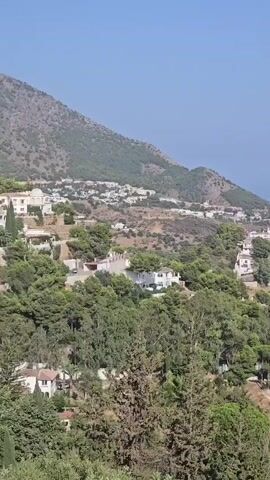 This is a panning drone shot of a Mediterranean coastal town. The camera moves from left to right, revealing a landscape dotted with white houses and lush green trees. In the distance, the blue sea meets the horizon under a clear sky. The terrain is hilly, with a mountain visible in the upper left corner. The shot captures the essence of a sunny day in a picturesque European settlement.