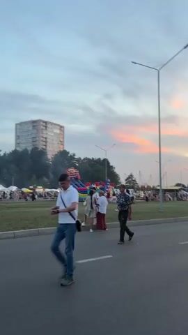 The video shows a street scene at dusk with a vibrant sunset in the background. There is a road in the foreground with a few people walking on it. To the left of the road is a grassy area with tents, stalls, and an inflatable playground. There are streetlights visible along the road. In the distance, there are trees and a tall building. The sky is filled with colorful clouds, with shades of orange, pink, and blue. The overall atmosphere appears to be that of an outdoor market or festival.