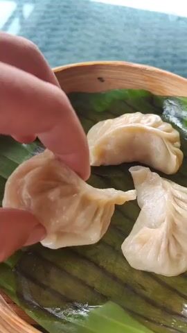 The video shows a close-up of a person's hand dipping a dim sum dumpling into a small bowl of orange sauce. The dumpling is light-colored and appears to be filled. It is being held between two fingers and thumb. In the background, there is a bamboo steamer basket with more dim sum inside, resting on a green leaf. A white plate is also visible in the background. The surface appears to be a table with a textured pattern. The lighting is bright and natural.