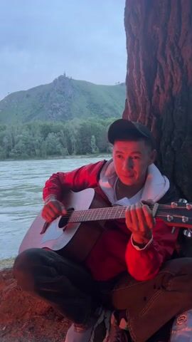 A man sits cross-legged under a tree by a river, playing an acoustic guitar. He wears a black baseball cap, a red and white jacket, dark pants, and dark shoes. The man strums the guitar and sings. The river flows behind him, with green hills and trees in the distance under a pale sky. The man's face and the guitar are illuminated by a warm orange light, likely from a campfire just out of frame.