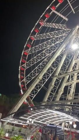 The video captures a close-up, upward-angled view of a large Ferris wheel at night. The Ferris wheel is illuminated with white lights forming a geometric pattern across its structure. Red gondolas are visible attached to the outer rim of the wheel, some of which are lit from within. A cluster of bright red lights is concentrated at the very top center of the wheel. Below the Ferris wheel, a covered area with transparent roofing and more lights is visible, with some indistinct figures of people and a sign that reads "WHEEL OF VENICE" in red lettering. The background is dark, indicating nighttime. The camera slowly pans and tilts, offering different perspectives of the Ferris wheel's rotating structure.