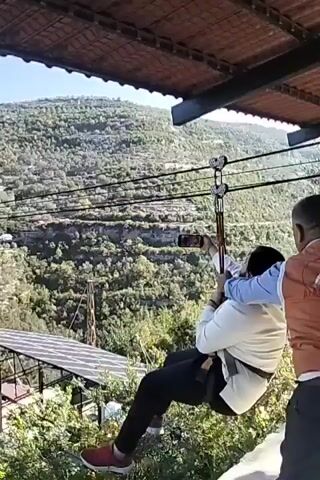 Here is a detailed description of the video:

A man on a zipline is moving from right to left in the video. He's wearing a white jacket, dark pants, and red shoes.  He appears to be holding a phone in his right hand, possibly recording his experience.

The zipline cable is visible, and it's anchored to a metal structure on either side of a green valley covered in trees and shrubs. The camera is positioned under a covered roof, framing the zipline and the man in the distance. The sky is clear and blue.

Another person wearing a blue sleeve is partly visible to the right of the person on the zipline.

A voice can be heard, saying "Bismillah Al-Atlal, Bravo Ahmad, on the wadi, not on your mobile, Bravo."