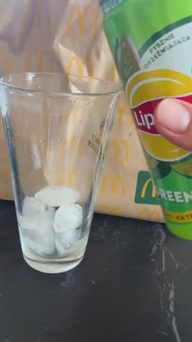 In this close-up shot, a hand is pouring a light-colored beverage from a green and white can into a clear glass filled with ice cubes. The can has text on it, and the glass has some writing and a small illustration on it. In the background, a brown paper bag with the McDonald's logo is visible. The scene is set on a dark, reflective surface, likely a countertop. The lighting suggests it could be daytime. The activity is the preparation of a cold drink, likely ice tea, as suggested by the text on the can. The overall impression is casual and domestic, possibly in a home kitchen.