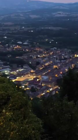 This video captures a nighttime aerial view of a town nestled in a valley. The town lights twinkle against the dark landscape, with rolling hills and distant mountains forming the backdrop. The sky is a dusky blue, transitioning into shades of pink and purple at the horizon, suggesting either twilight or dawn. The foreground is dominated by dark, shadowy foliage, likely trees, that partially obscure the view of the town. The overall impression is one of a peaceful, illuminated settlement in a rural, mountainous region.