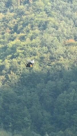 The video shows a person riding a zipline. The zipline is a cable stretched across a forested area. The person is wearing a helmet and is attached to the zipline by a harness. The person is moving from left to right in the frame. The forest is dense and green. The sky is not visible. The video is shaky and the focus is not perfect.