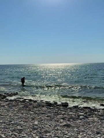 This is a vertical video of a rocky beach on a sunny day. The sun is high in the sky, creating a bright glare on the surface of the calm, blue sea. Gentle waves lap against the shore, which is covered in smooth, grey and white pebbles. In the distance, the vast expanse of the ocean meets the clear blue sky, with a faint landmass visible on the horizon.

A lone figure, silhouetted against the bright water, stands partially submerged in the shallow waves, facing away from the camera. They appear to be wearing dark clothing and possibly a backpack. The overall mood is peaceful and serene, suggesting a quiet moment of contemplation or enjoyment of nature. The scene evokes a sense of summer tranquility and the simple beauty of a coastal landscape. There is no visible text in the video.