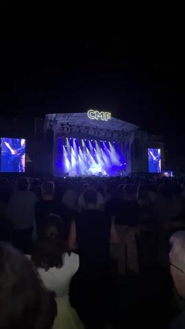 The video shows a night concert with a large crowd facing a brightly lit stage. The stage has a banner with "CMF" in large neon letters. Blue and white spotlights are directed at the stage. There are large screens on either side of the stage showing close-ups of performers. The crowd is diverse in age and gender, with many people standing and facing the stage. Some people are holding up their phones, likely to record the performance. The overall atmosphere appears to be energetic and festive.