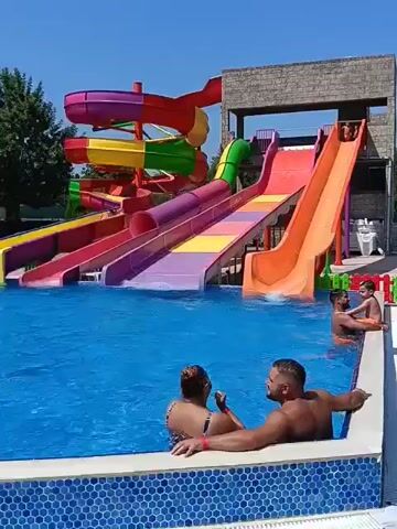 The video opens with a wide shot of a water park on a clear, sunny day. In the foreground, two adults are partially submerged in a bright blue swimming pool, facing away from the camera. The man on the right has his arm around the woman's shoulder. In the background, a large, multi-colored water slide structure dominates the scene. The slides are a vibrant mix of purple, pink, yellow, green, and orange, with a red spiraling section at the top. Two orange slides and a red slide are visible side-by-side, with people on them. One person is seen at the top of the orange slide on the right, about to descend, and another person is at the bottom of the same slide. Two children are in the pool near the orange slide. The pool area has a tiled edge. To the right of the slides, a building with a stone-like facade stands, and beyond that, a fence with colorful posts. Trees are visible behind the water slide structure. The video appears to be a short, unedited clip, possibly from a personal recording, showing a recreational activity at a water park.