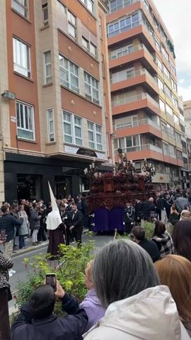 The video shows a religious procession taking place on a city street lined with apartment buildings. A large, elaborately decorated float, featuring a statue of Jesus Christ, is being carried through a crowd of onlookers. The float is adorned with candles, flowers, and intricate wooden carvings, and is draped in a purple cloth. People are standing on both sides of the street, watching and recording the procession with their phones. Some individuals in the procession are wearing traditional religious garb, including tall, pointed hoods.  The weather appears overcast. A business named "Liberty Horizon" is visible in the background.