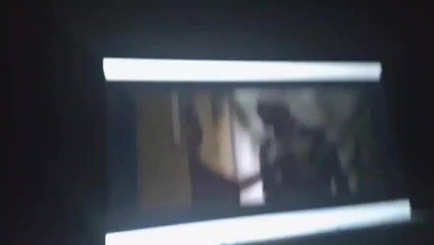 The video displays a scene from a film or television show, projected onto a screen. The frame shows two individuals, both appearing to be adult males, on a motorcycle. The man in the front is wearing a plaid shirt and a white bandage around his head. The man behind him is wearing a striped shirt and also appears to have a bandage on his head. The motorcycle is moving, as indicated by the motion blur in the background. The scene appears to be set outdoors, possibly in a rural or semi-urban area, with some greenery and a building visible behind the subjects. The lighting suggests it could be daytime, but the overall image quality is poor, making it difficult to ascertain specific details about the environment or the time of day. There is no visible text in the frame. The interaction between the two individuals is unclear due to the quality and the angle of the shot, but their presence on a motorcycle suggests they are traveling or involved in some form of action sequence.