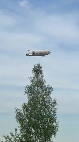 A white zeppelin labeled "Europa Park" floats in the sky above a tree. The sky is light blue with some clouds. A cartoon character is depicted on the side of the zeppelin.