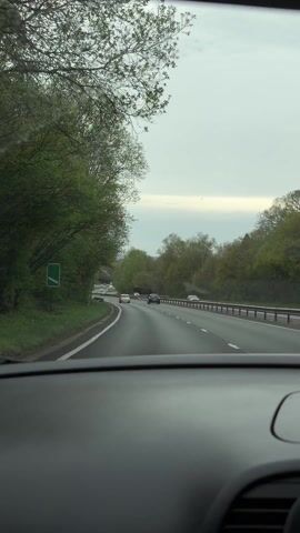 A car is driving on a highway. The windshield is in focus. There are other cars ahead on the highway. There is a sign indicating exits to Portsmouth, Petersfield, and Liphook. Trees line either side of the road. The sky is overcast.