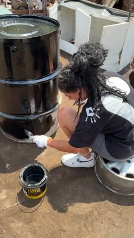 A person wearing a black t-shirt and grey shorts is crouched down next to a large, black, cylindrical object. They are wearing white gloves and holding a paintbrush, which they are using to apply black paint to the bottom section of the cylinder. A can of black paint sits on the ground next to them. The person has long, dark hair pulled up into a bun. They are sitting on a silver car wheel. The background shows a cluttered outdoor area with various metal objects and what appears to be a white cabinet. The ground is covered in dirt and gravel. Sunlight is casting shadows.