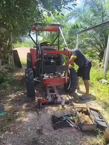 A man is working on the back of a red tractor. He is wearing a dark shirt and shorts. The tractor is on a lift. There are tools scattered on the ground around the tractor. The scene appears to be outdoors, with trees and palm trees in the background. The ground is dirt and gravel. The lighting suggests it is daytime.