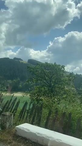 The video shows a view from inside a vehicle, likely a car, moving along a road. The scene outside the window is dominated by lush green trees and rolling hills covered in dense forests. In the distance, a body of water, possibly a lake or reservoir, with a greenish hue is visible, partially obscured by trees and a fence. The sky is partly cloudy with large white cumulus clouds against a blue background. Power lines are visible stretching across the top of the frame. In the foreground, parts of the vehicle, including a guardrail and window reflections, are present. One segment of the video shows a person wearing a patterned shirt and dark pants, partially visible behind some foliage. The overall impression is of a scenic drive through a mountainous, forested region on a bright, cloudy day.